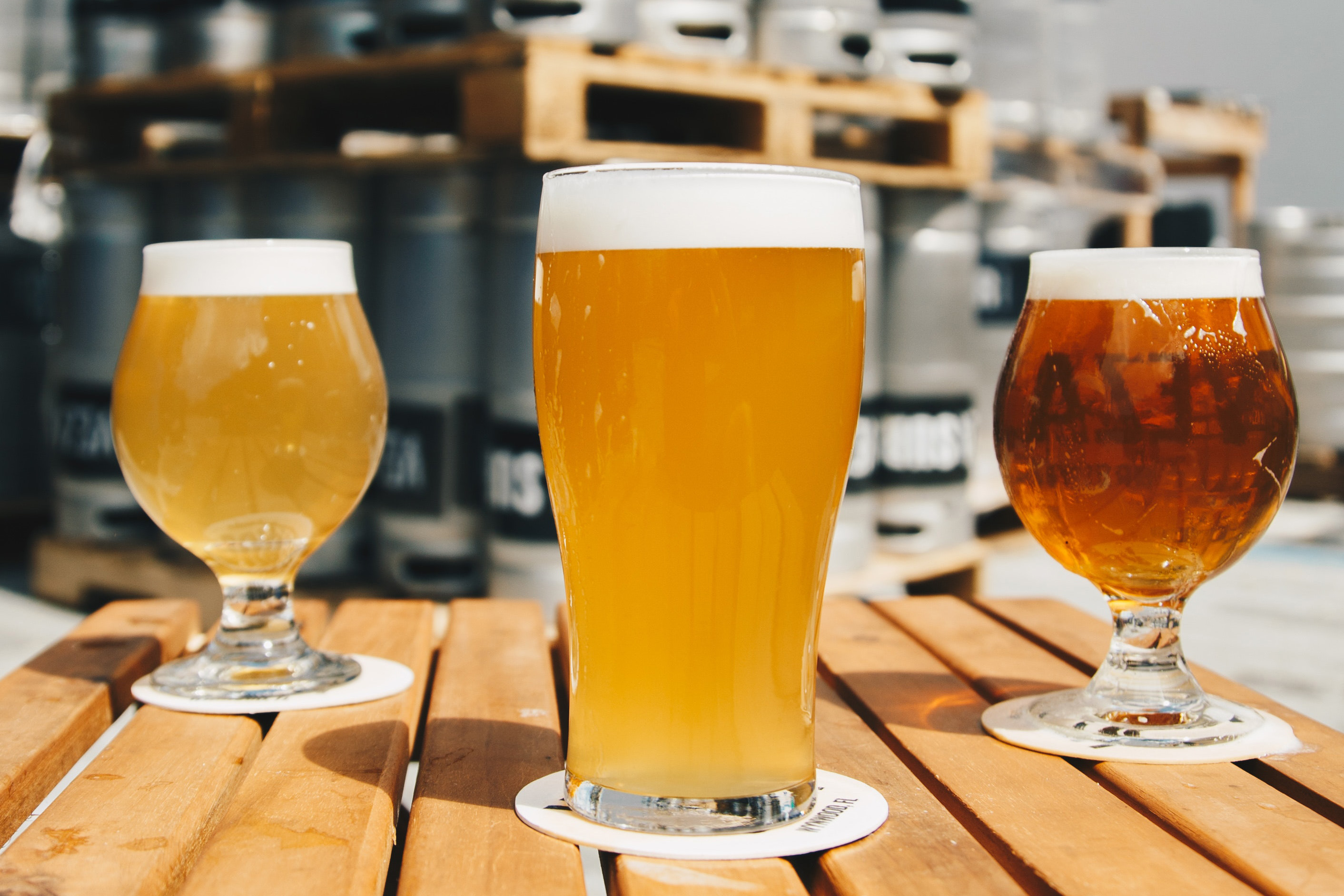 3 glasses of beer sitting on a table in front of lots of beer kegs.
